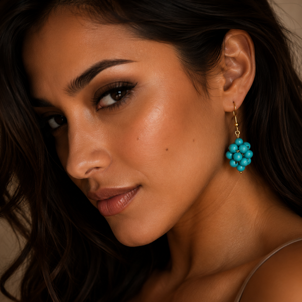 A close-up lifestyle shot of a woman wearing a gold dangle earring with a cluster of turquoise blue beads, set against a warm beige background.