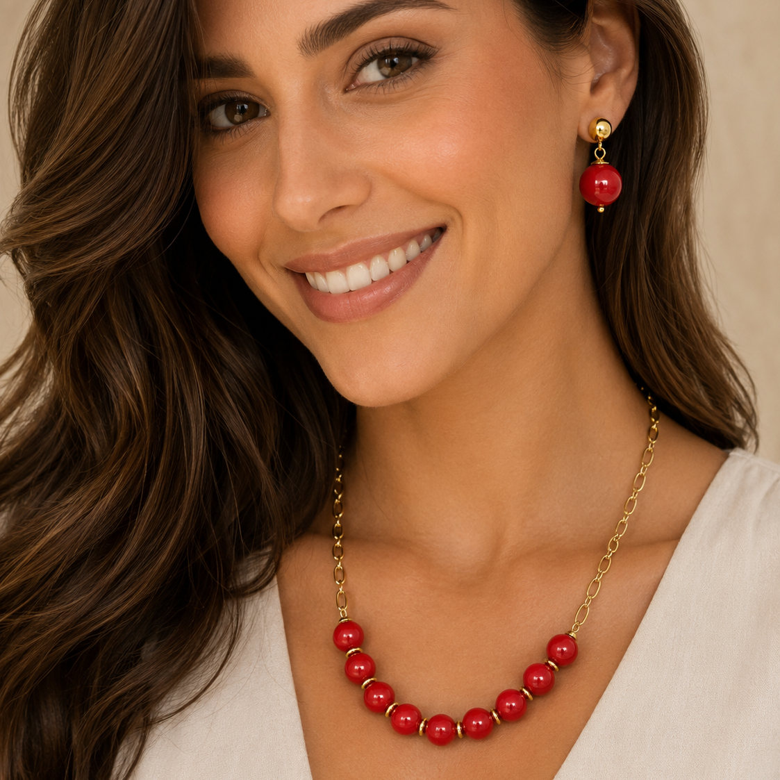 A smiling woman with long brown hair models a gold chain necklace and matching drop earring featuring glossy red beads, set against a soft beige background.