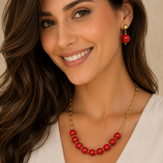A smiling woman with long brown hair models a gold chain necklace and matching drop earring featuring glossy red beads, set against a soft beige background.