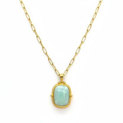 A gold pendant necklace with a light green stone, displayed on a textured white jewelry cushion.