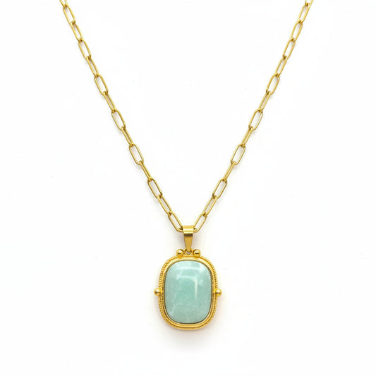 A gold pendant necklace with a light green stone, displayed on a textured white jewelry cushion.