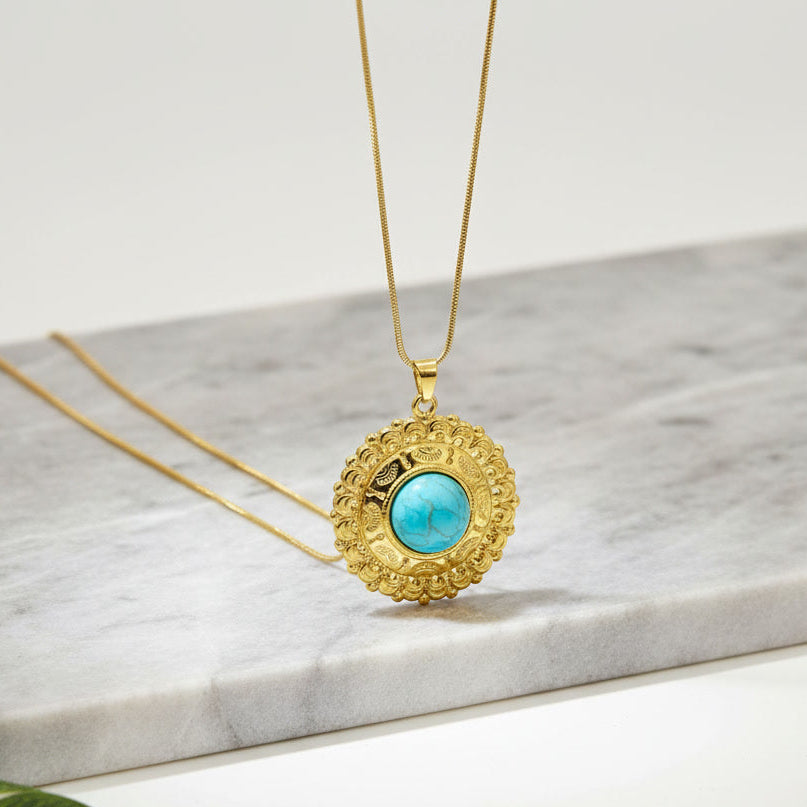 A gold pendant necklace with a textured circular frame and a central turquoise-blue stone, displayed on a white marble slab with a green leaf in the corner.